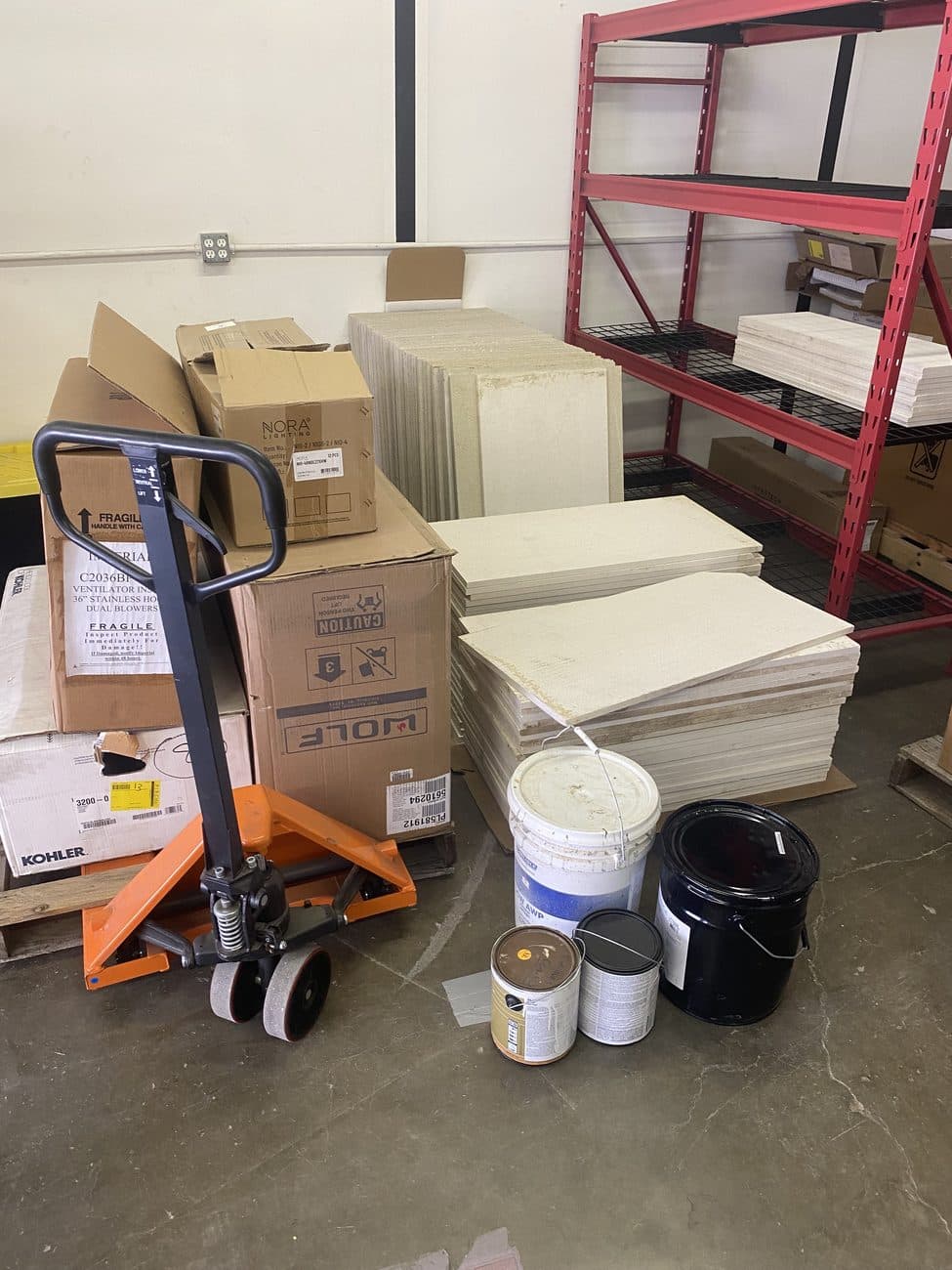 Mixed surplus construction materials in a warehouse — palletised stone, boxed fixtures and paint, with a pallet jack alongside